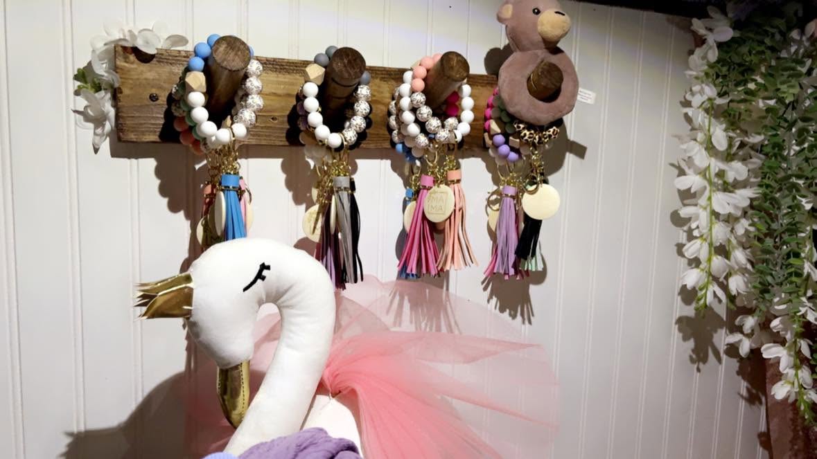 Beaded keychains with tassels hanging on a reclaimed wood rack beside silk florals.